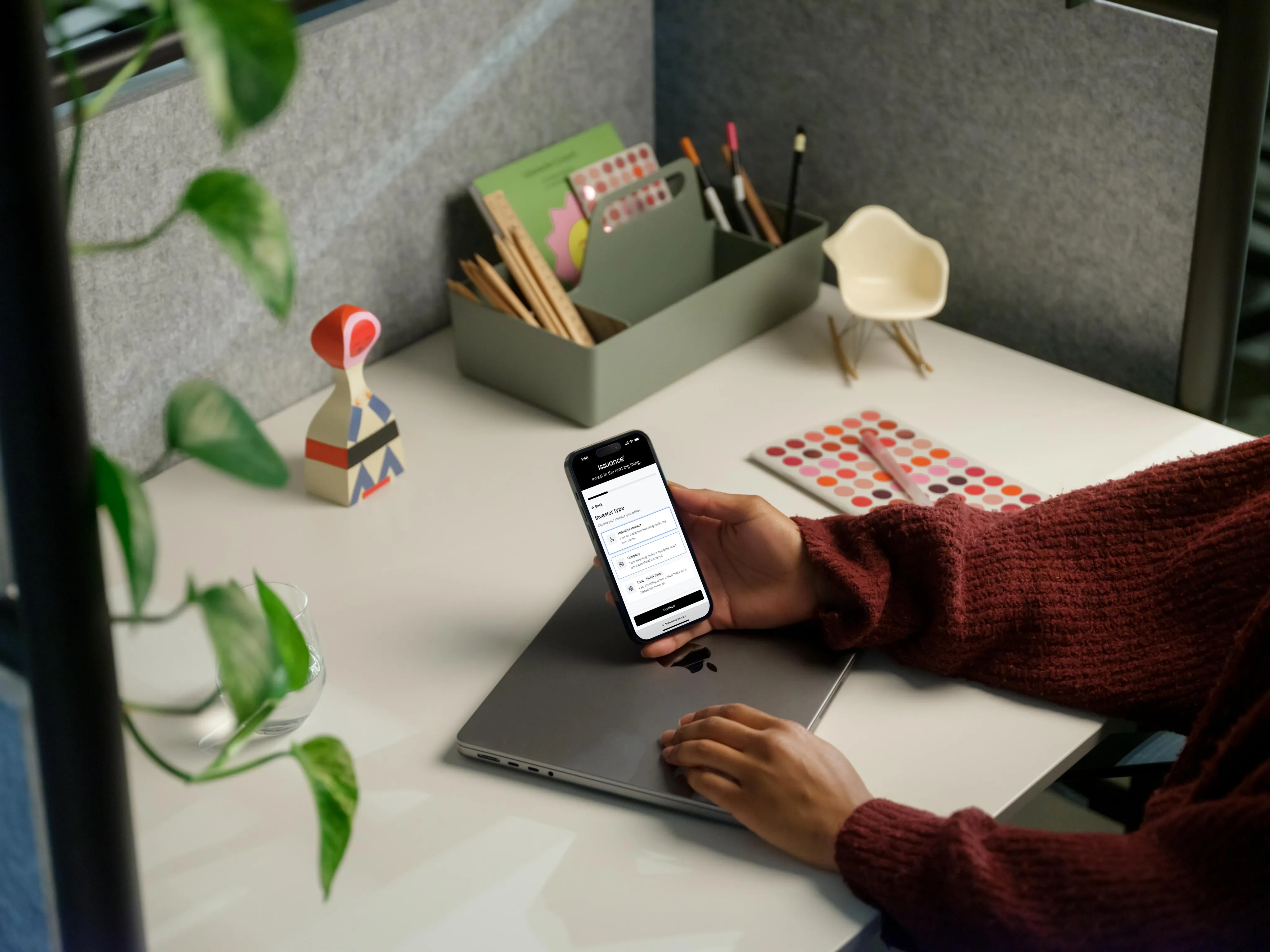 A person at a desk holding a phone that shows the Issuance investor flow, open at the investor type step